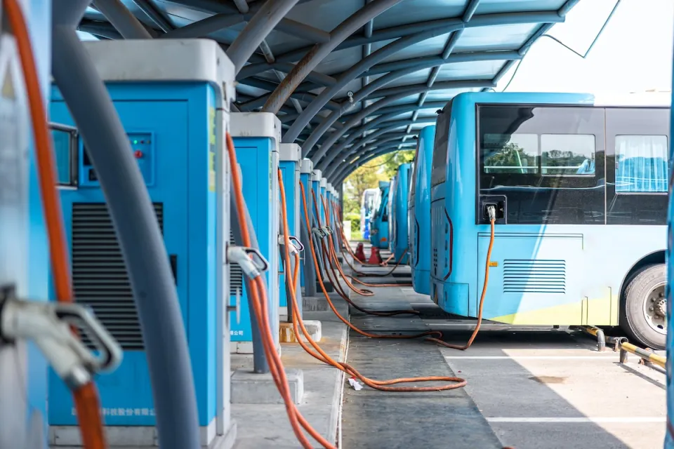 Row of blue electric buses parked and plugged in at charging station.
